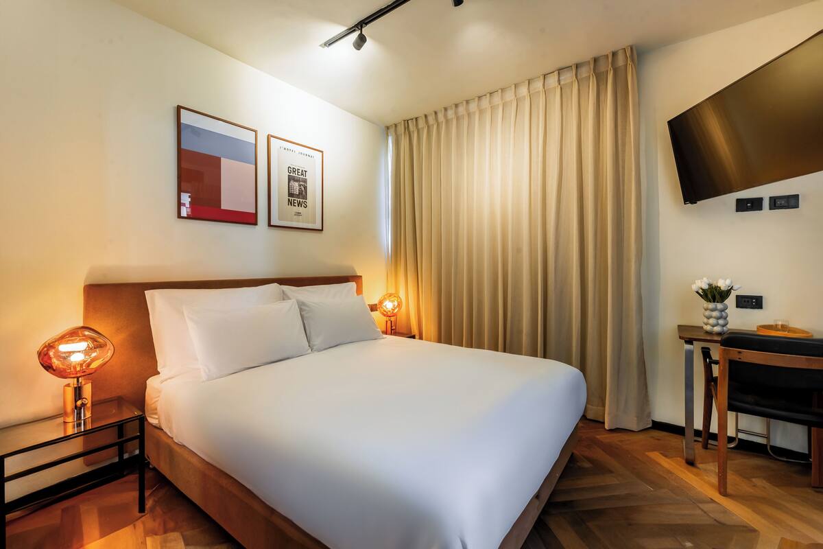 Een hotelkamer met een bed, bedlampen, een flatscreen televisie, een kleintje tafeltje met een vaas, en gordijnen.