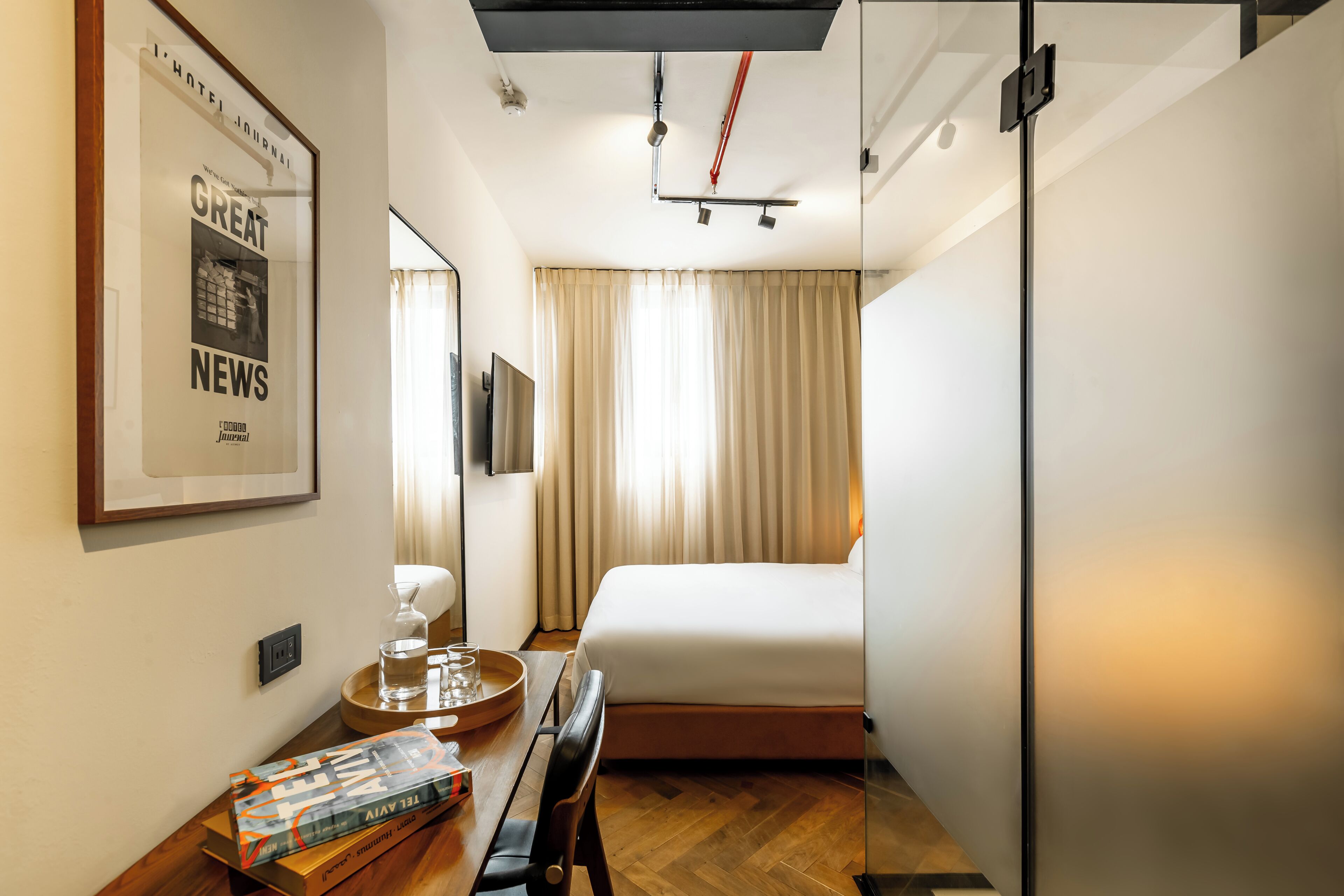 A hotel room with a bed, a desk with a tray, a glass of water, a framed picture on the wall, and a window with curtains.