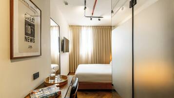 A hotel room with a bed, a desk with a tray, a glass of water, a framed picture on the wall, and a window with curtains.