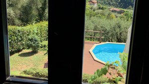 Vista dalla finestra su una piscina, circondata da vegetazione lussureggiante e dolci colline in lontananza.