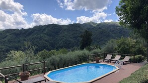 Una piscina rettangolare con bordo in legno e poltrone prendisole, circondata da una recinzione in legno, con montagne sullo sfondo.