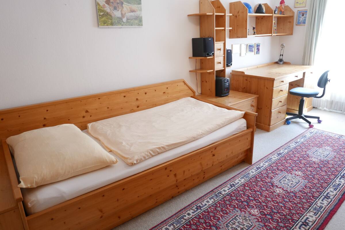 En säng av trä med en madrass och kuddar, ett skrivbord med en stol, en hylla med diverse föremål och en matta.