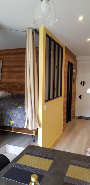 Een moderne slaapkamer met een houten hoofdbord, een bed met een bloemenbedekking, een geel accentmuur en een groot raam.