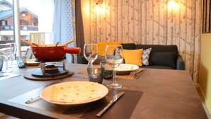 A dining table set with plates, cutlery, and glasses, a red pot on a stand, and a sofa with cushions in a room with patterned wallpaper and a window view. - Elegant and modern, at the foot of Mt-Blanc (Saint-Gervais-les-Bains)