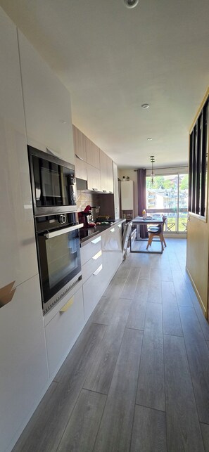 Een moderne keuken met inbouwapparatuur, een eethoek met een tafel en stoelen, en uitzicht op buiten door grote ramen.