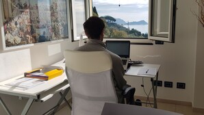 A person seated at a desk with a laptop, papers, and a pen, in a room with a large window overlooking a scenic landscape. - Maiso house nestled between the sea and olive trees (Lavagna)