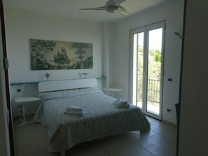 Une chambre à coucher comprenant un lit, une table de chevet, un tableau, un ventilateur de plafond et une porte-fenêtre coulissante donnant sur un balcon.