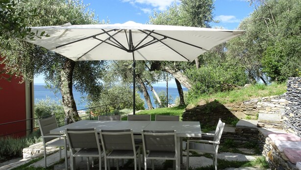 An outdoor dining area with a table and chairs under a large umbrella, overlooking a body of water. - Maiso house nestled between the sea and olive trees (Lavagna)