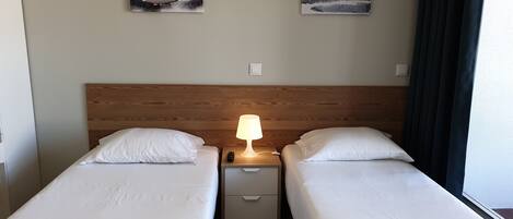 A hotel room with two single beds, a wooden headboard, a nightstand with a lamp, and two wall-mounted paintings of sailboats.