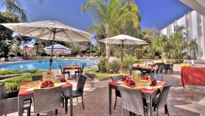 Un área de comedor al aire libre con mesas y sillas, sombrillas y una piscina al fondo.