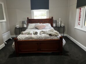 A neatly made bed with a floral-patterned bedspread, two bedside tables with lamps, a window with a dark blind, and a radiator. - Skibo Cottage (Skibo)