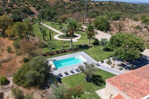 A swimming pool with a surrounding deck, a stone building with a tiled roof, and a landscaped garden with trees and shrubs.
