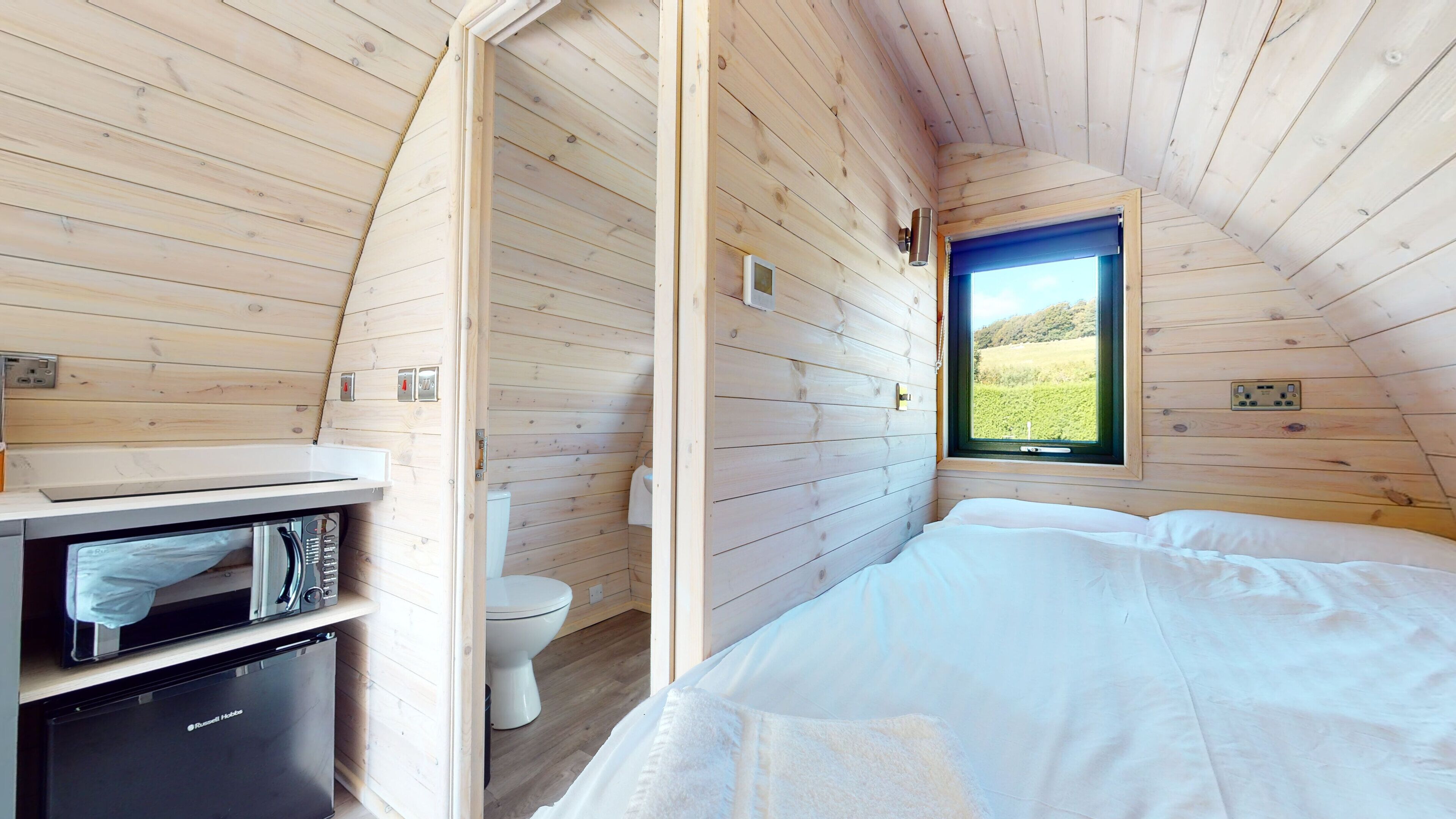 A compact wooden cabin room with a bed, a small kitchenette with a refrigerator, a toilet, and a window with a view of a green landscape.