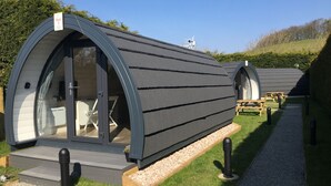 Il s'agit d'un hébergement de type « capsule », à l'extérieur incurvé, avec un intérieur visible comprenant un lit et un coin salon, et un sentier de gravier menant à des tables de pique-nique.