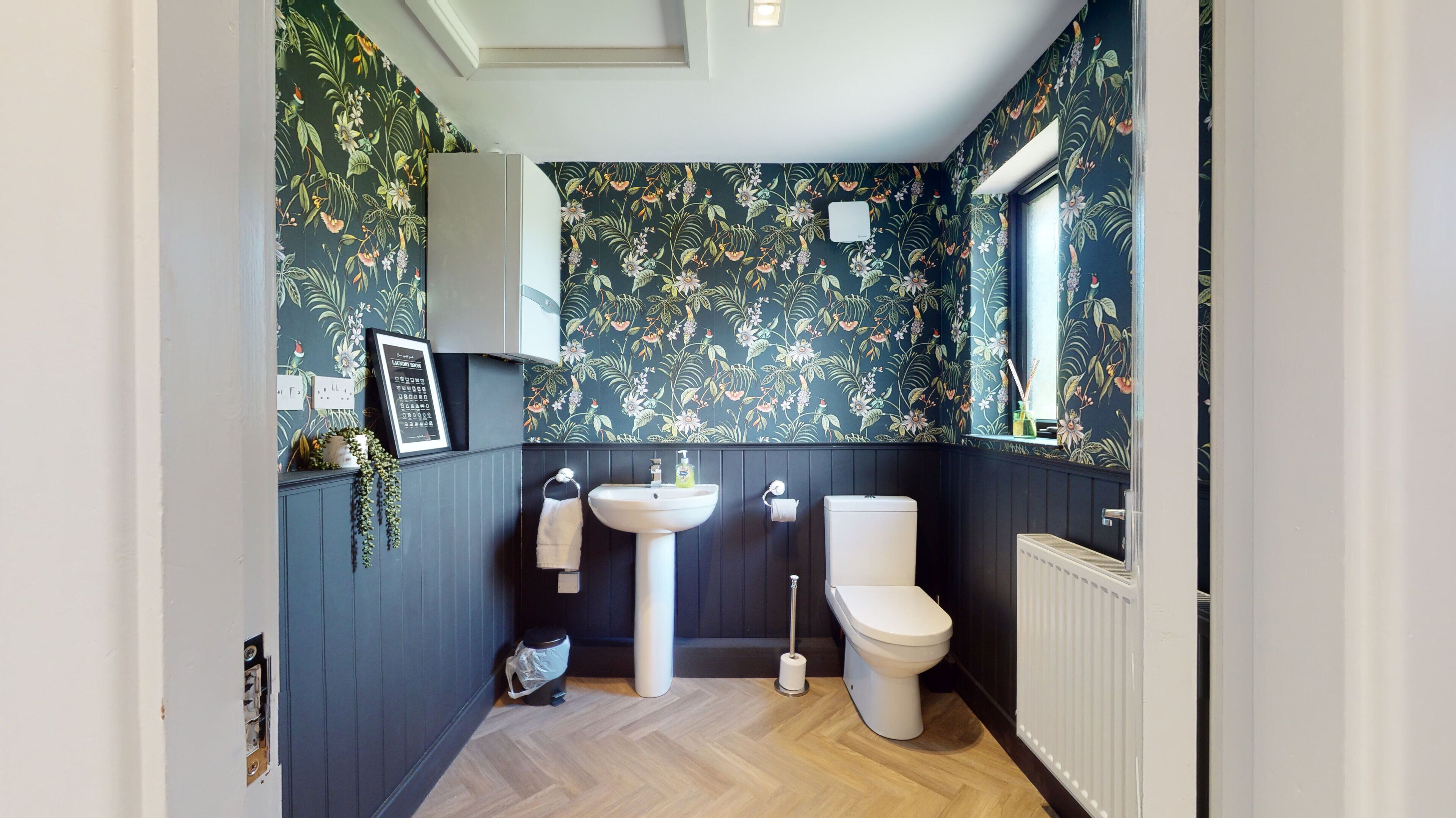 Ein Badezimmer mit dunkel gestreifter Blüten-Tapete, einem weißen Waschbecken, einer Toilette und einem Heizkörper.
