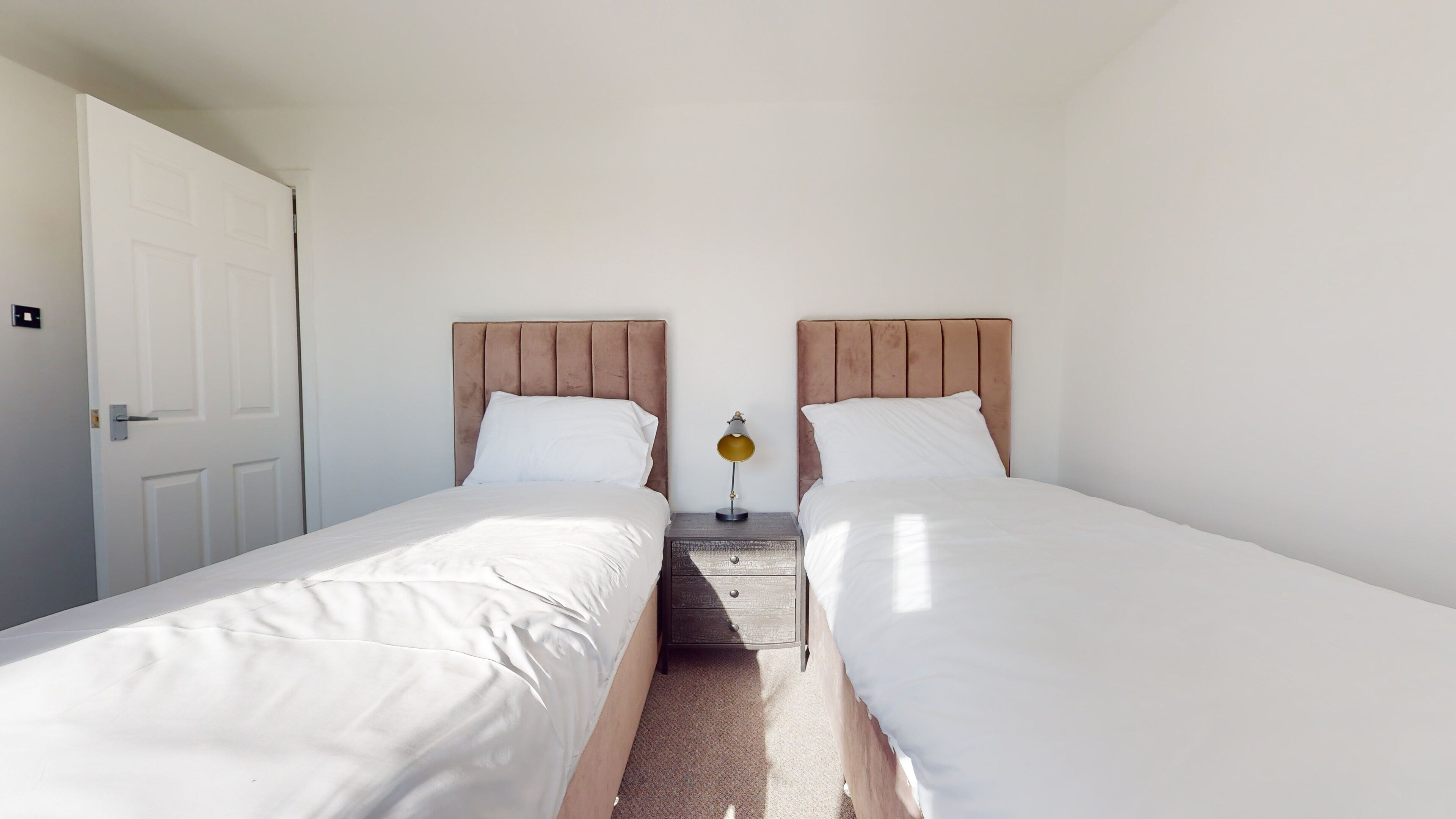 A twin bedroom with two single beds, a nightstand with a lamp, and a white door.