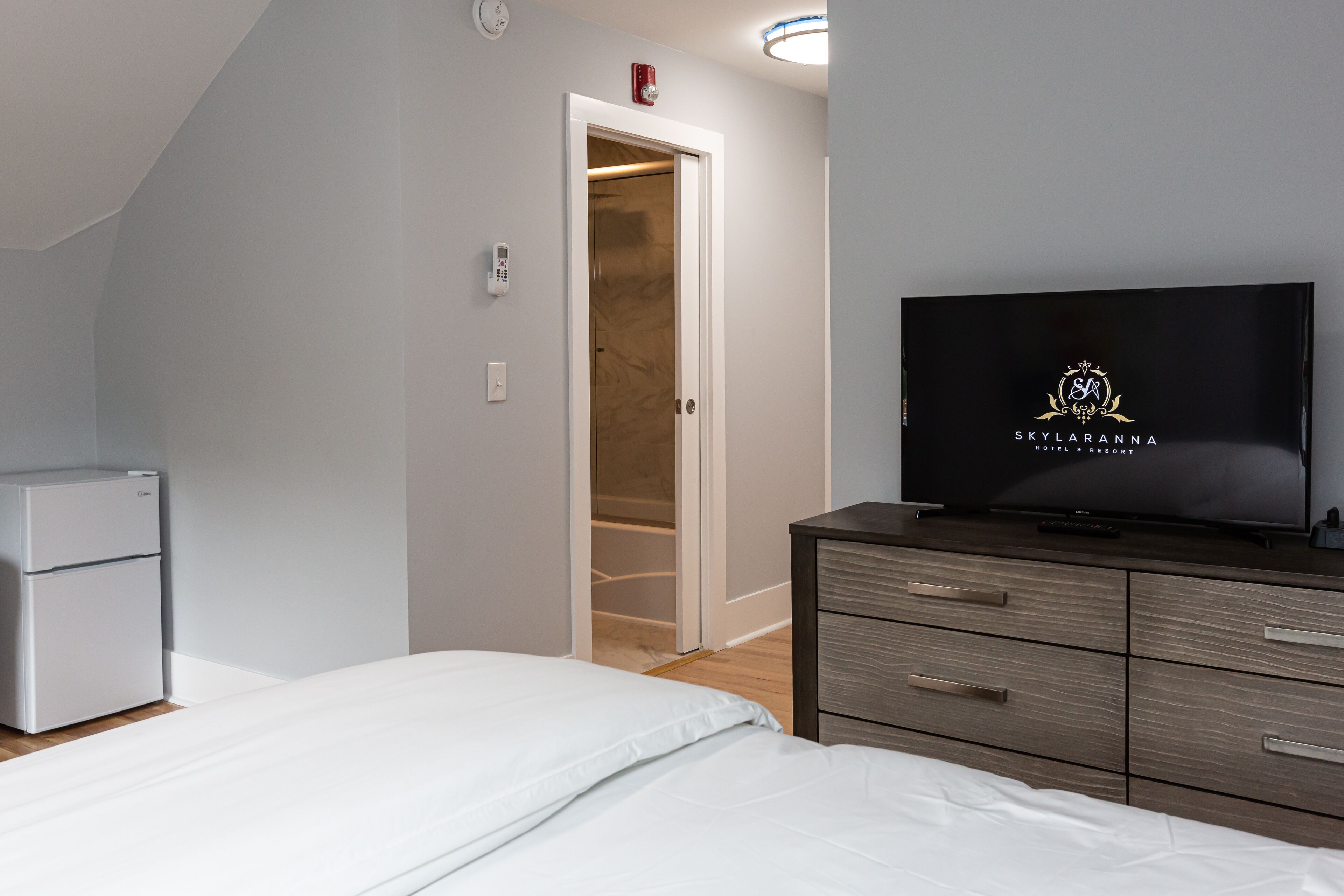 Un dormitorio con cama, televisión, armario, refrigerador pequeño y puerta que da a un baño.