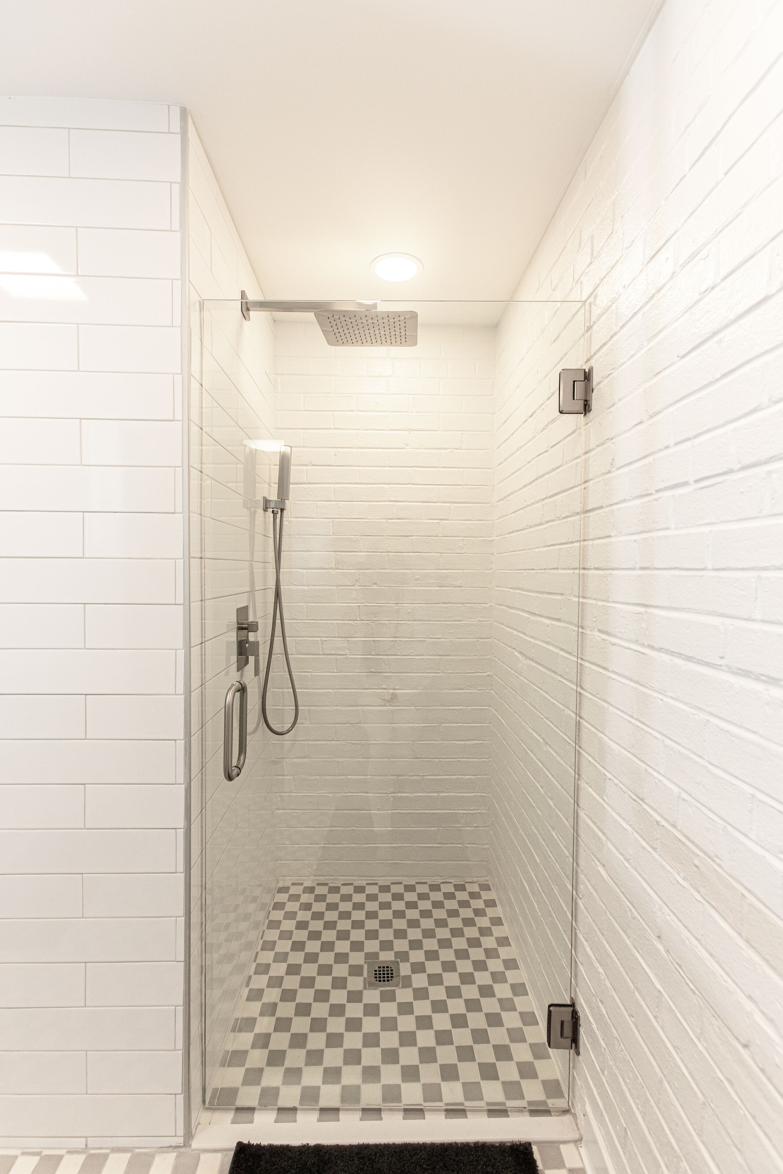 Una ducha con mampara de vidrio, suelo de cuadros, paredes de azulejos blancos y desagüe visible.