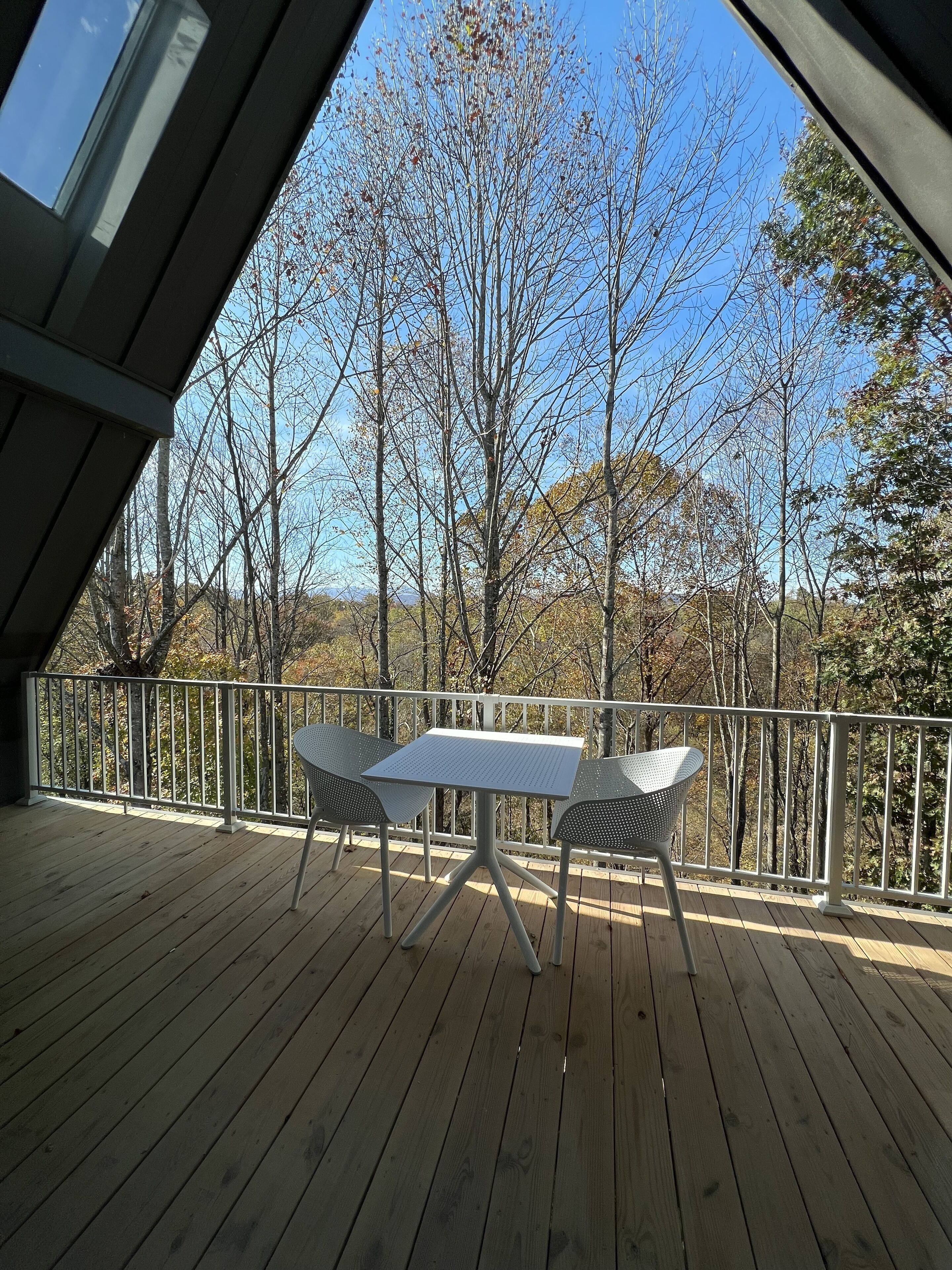 A wooden deck with a table and chairs, surrounded by a white railing, overlooking a forested area.