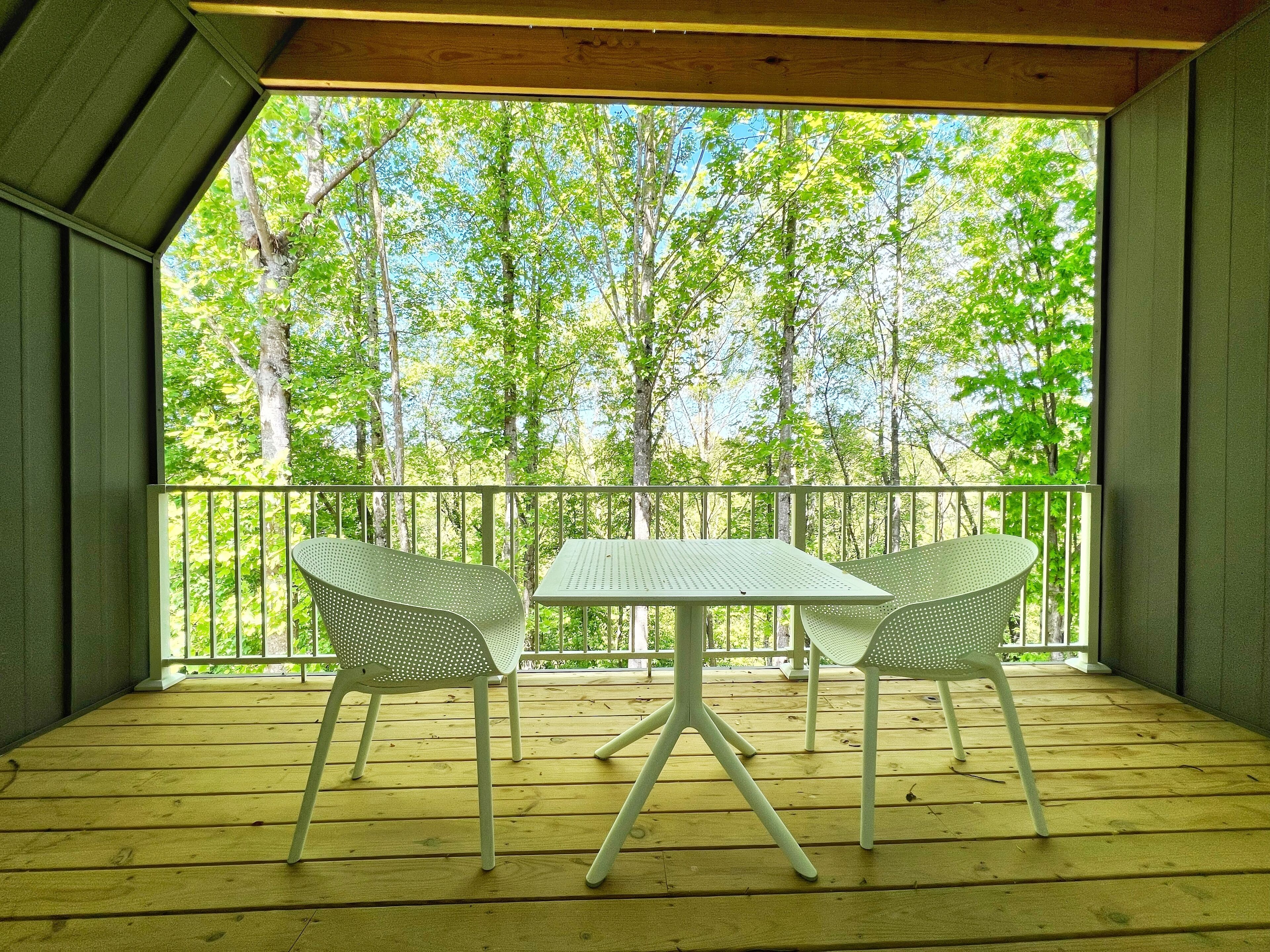 Um deck de madeira com uma mesa e duas cadeiras, com vista para uma área arborizada.