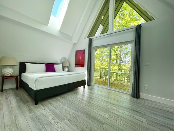 Un dormitorio con cama, mesita de noche, lámpara y vistas a un balcón con mobiliario exterior.