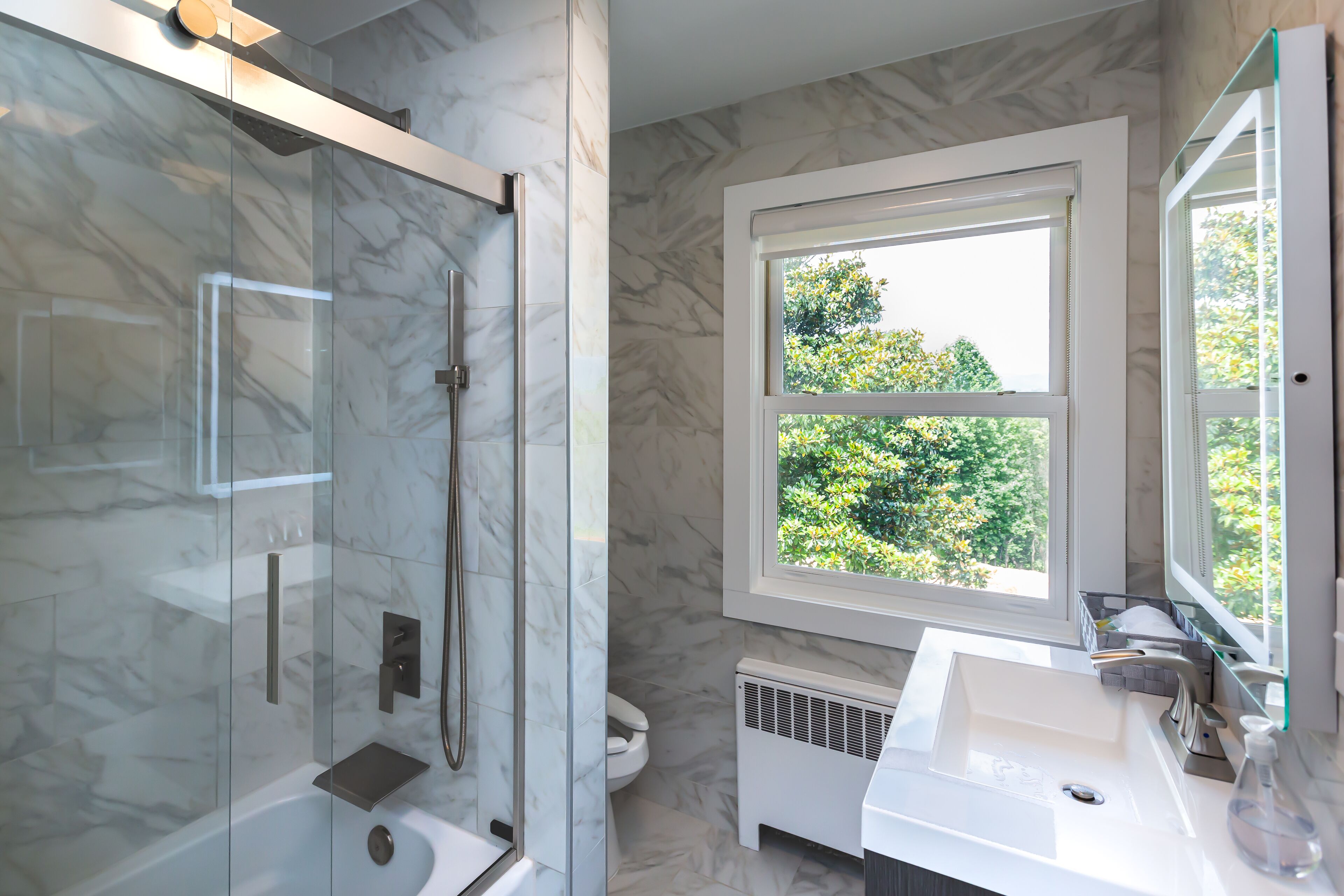 Un baño moderno con ducha de vidrio, lavamanos blanco y ventana con vista a la vegetación.