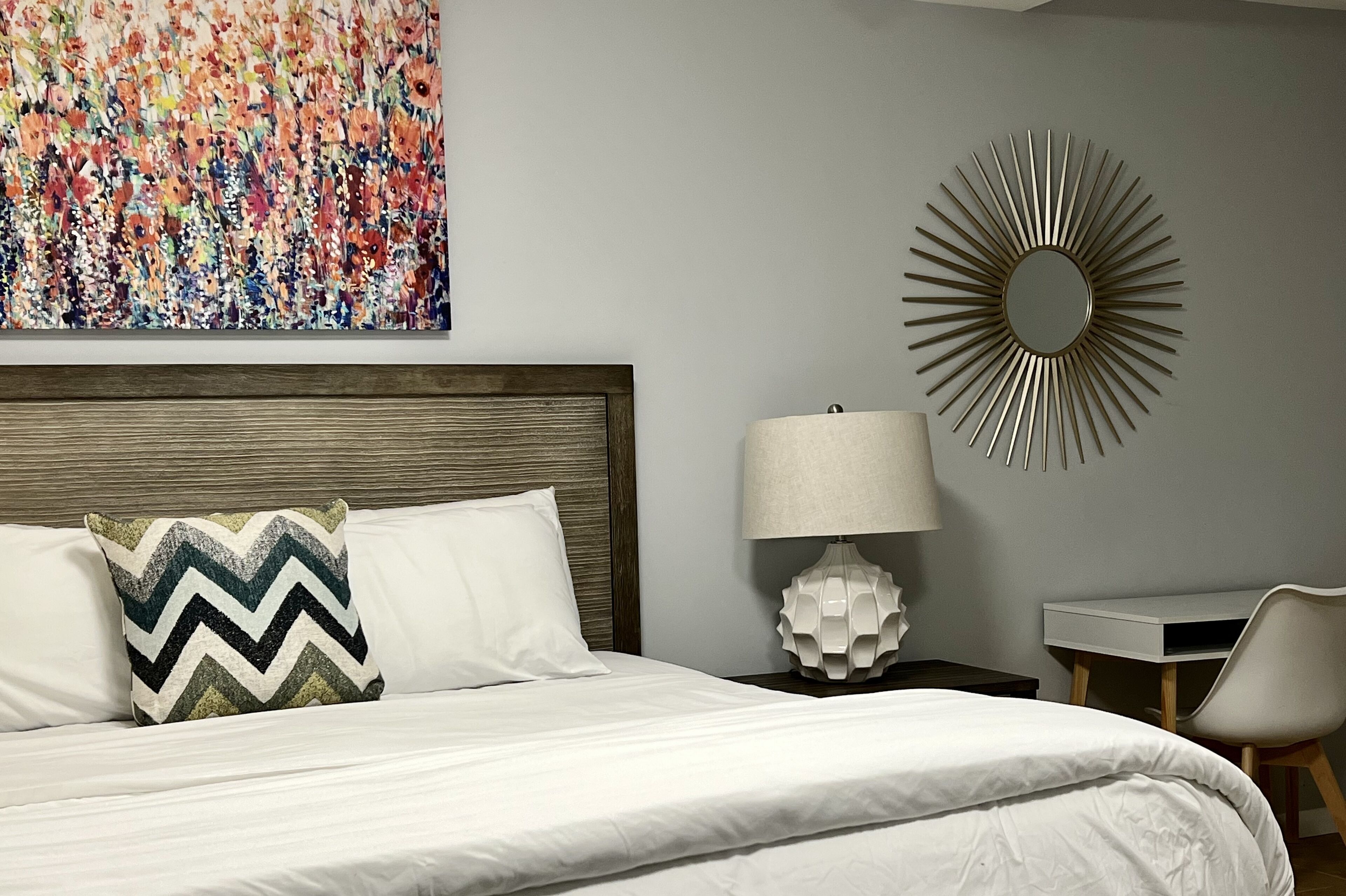 Una cama bien hecha con un almohadón de estampado en zigzag, un espejo redondo, un escritorio con una silla y un cuadro abstracto colorido en la pared.
