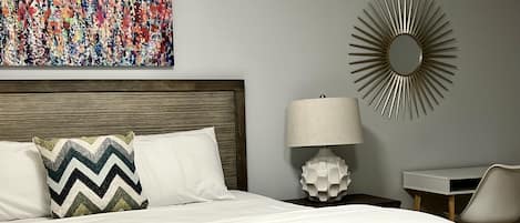 Una cama bien hecha con un almohadón de estampado en zigzag, un espejo redondo, un escritorio con una silla y un cuadro abstracto colorido en la pared.