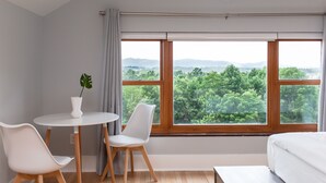 Un dormitorio con un ventanal grande, una cama, una mesita con sillas y una planta en maceta.