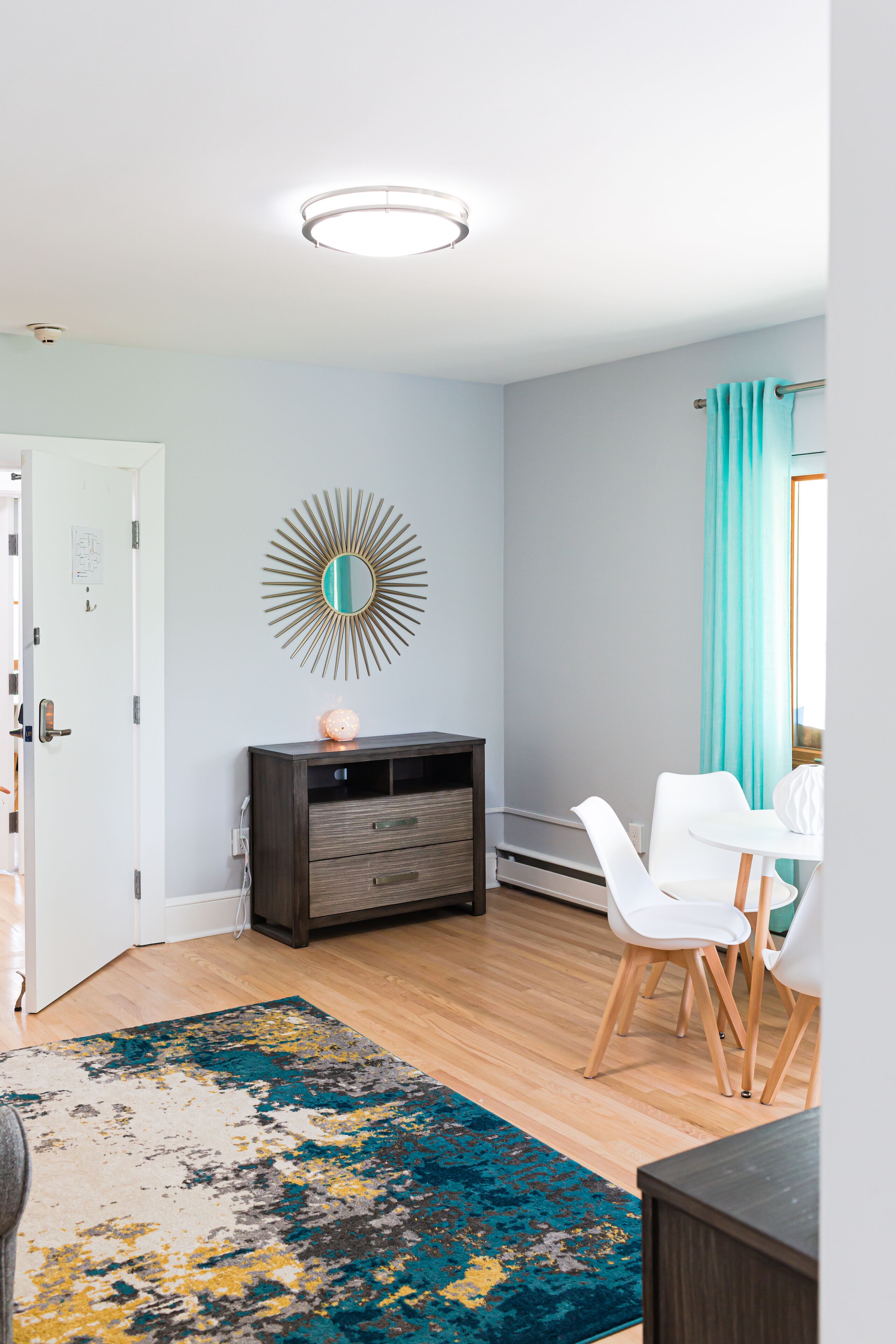 A modern living room with a wooden floor, a dark-colored cabinet, a white chair, a round mirror, and a teal curtain.