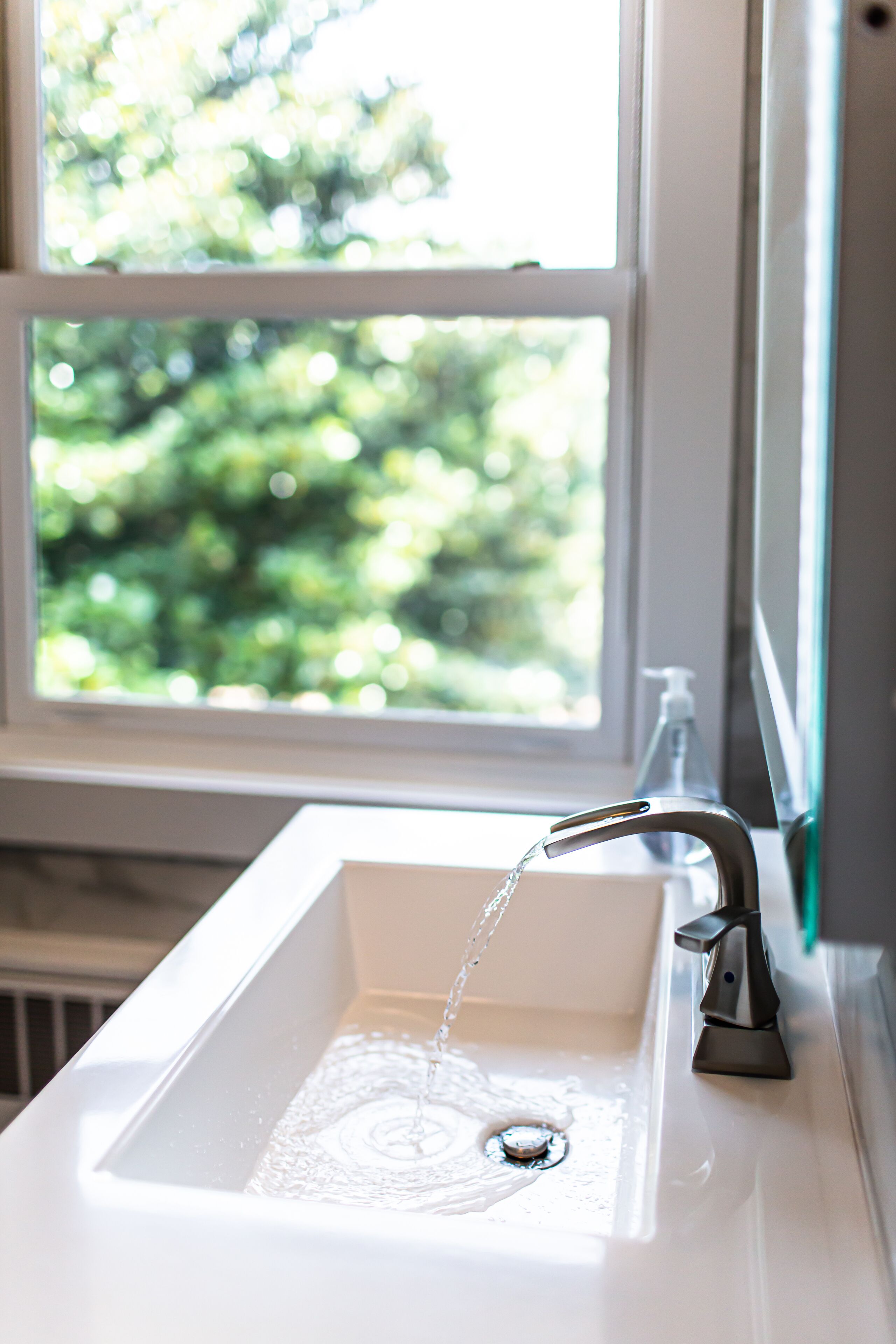 A bathroom sink with running water, a window, and a view of greenery outside.