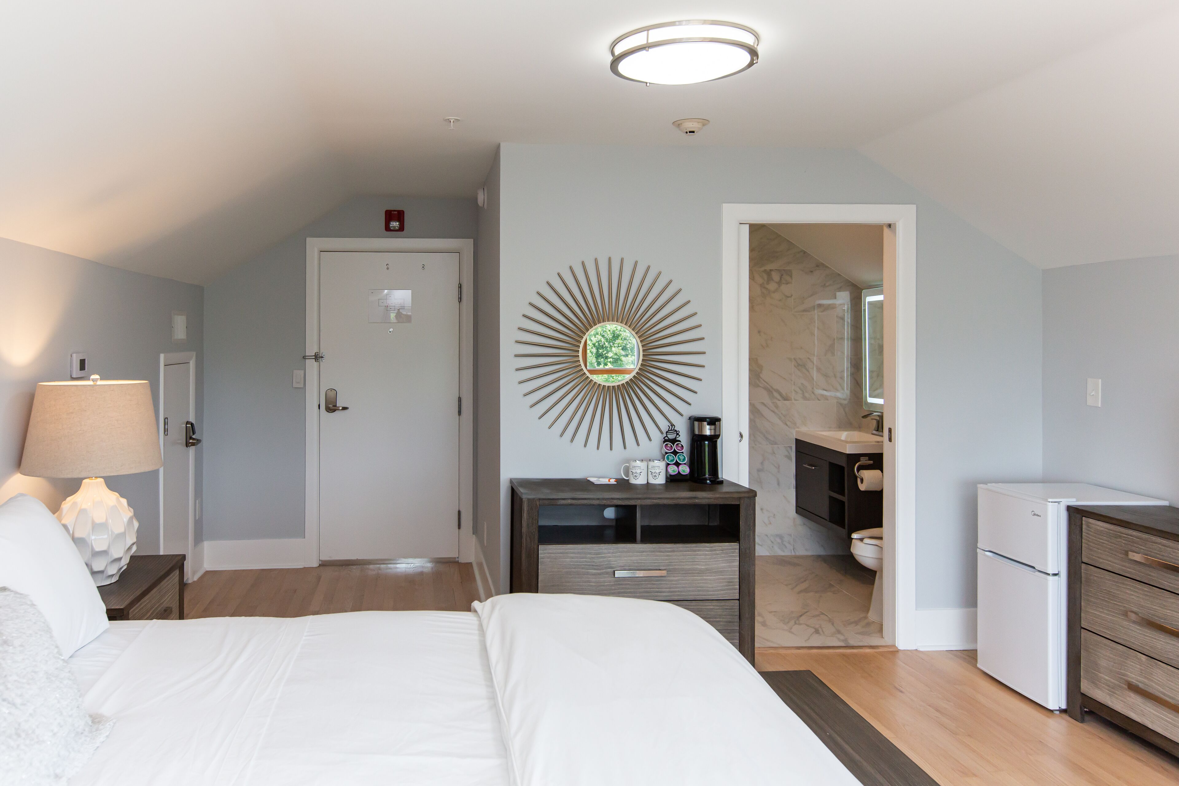 Habitación de hotel con cama, mesita de noche, lámpara, decoración en la pared y una zona de kitchenette.