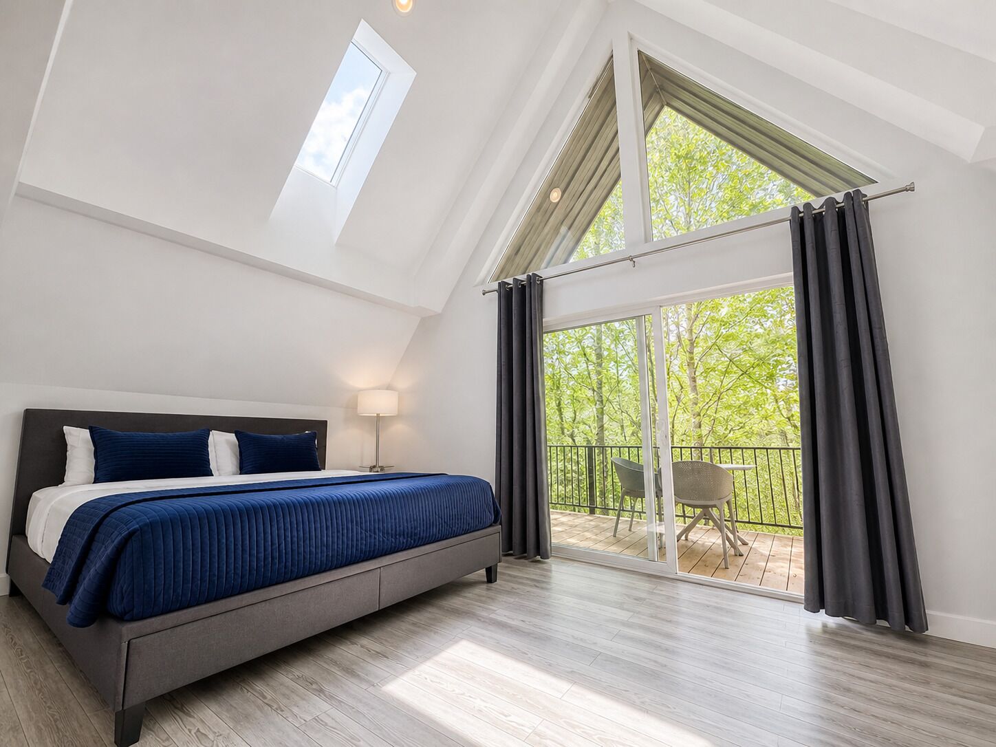 A bedroom with a large bed, a nightstand, a lamp, and a view of a balcony with outdoor furniture.