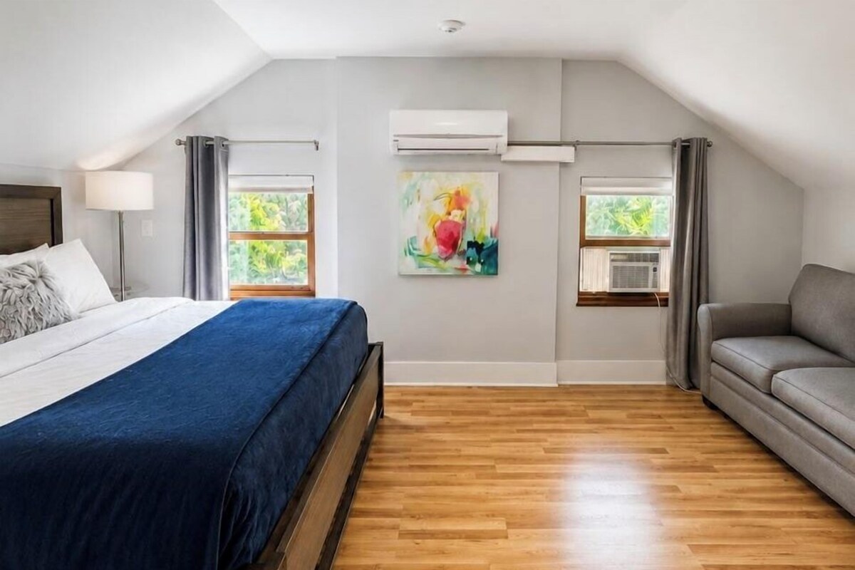 Un dormitorio con cama, sofá, dos ventanas con cortinas, un equipo de aire acondicionado y un cuadro en la pared.