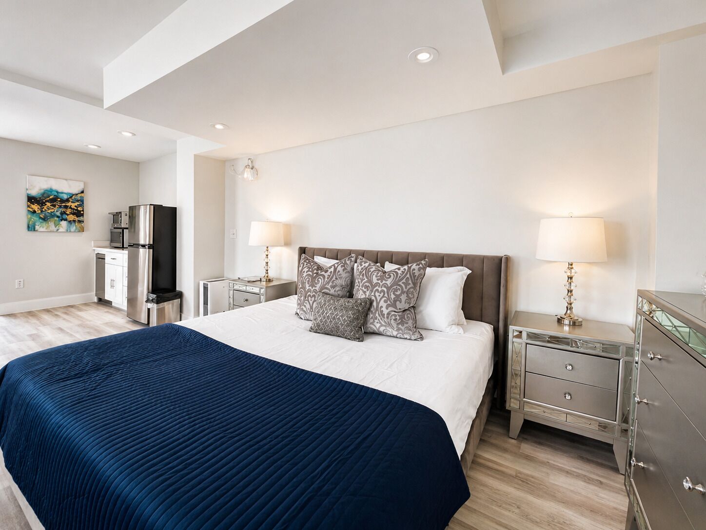 A modern hotel room with a large bed, bedside tables with lamps, a refrigerator, and a microwave in the kitchenette area.