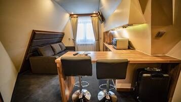 Habitación de hotel compacta con kitchenette, una zona de comedor con dos taburetes tipo bar, un sofá y una maleta grande.