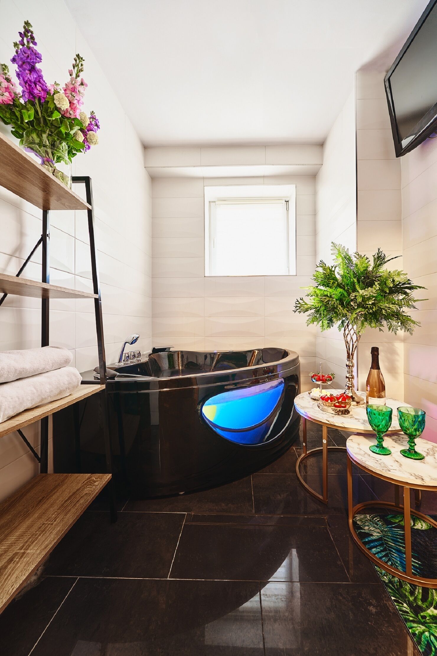 A modern bathroom with a freestanding bathtub, a wooden bench, a small table with decorative items, and a window providing natural light.