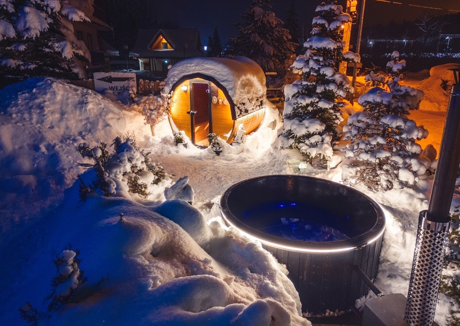 Zona exterior cubierta de nieve con una estructura de madera que incluye una entrada circular, un jacuzzi y un cartel con flechas direccionales.