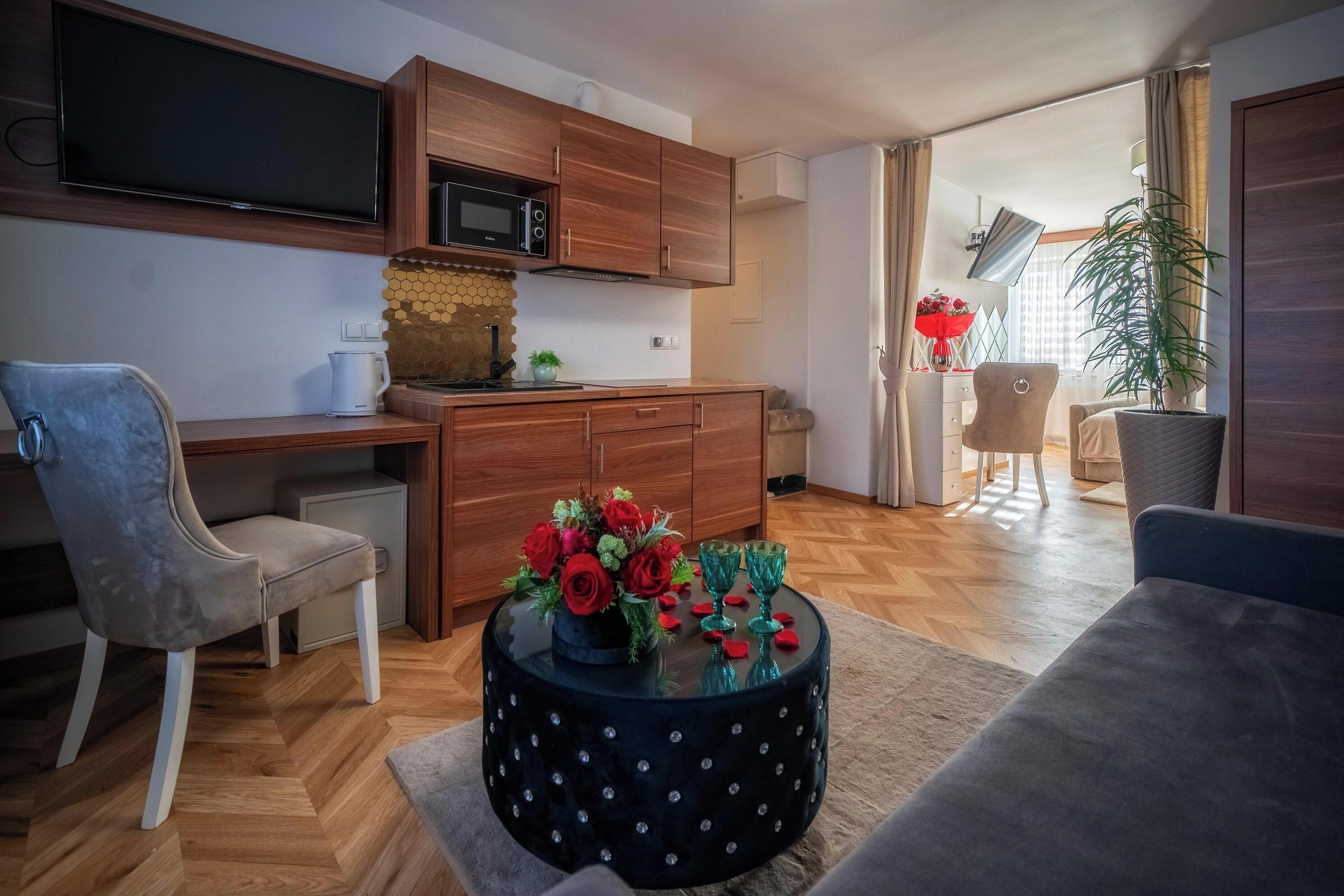 A modern living room with a sofa, a coffee table with a floral centerpiece, a television mounted above a wooden cabinet, and a kitchen area with a microwave.