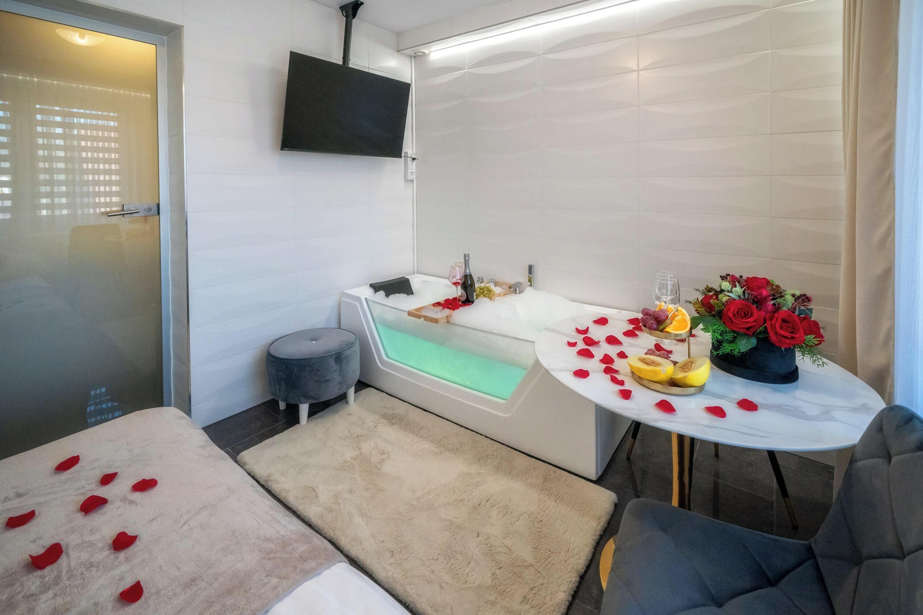 Un baño moderno con bañera, una mesita redonda con pétalos de rosa y fruta, y un televisor montado en la pared.