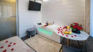 Un baño moderno con bañera, una mesita redonda con pétalos de rosa y fruta, y un televisor montado en la pared.