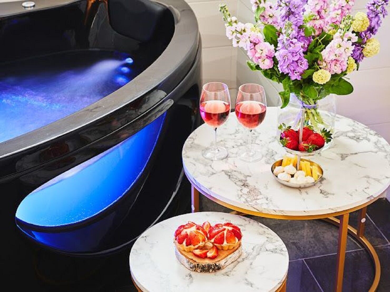 A spa area with a hot tub, two glasses of red wine, a vase with flowers, and a plate of fruit.