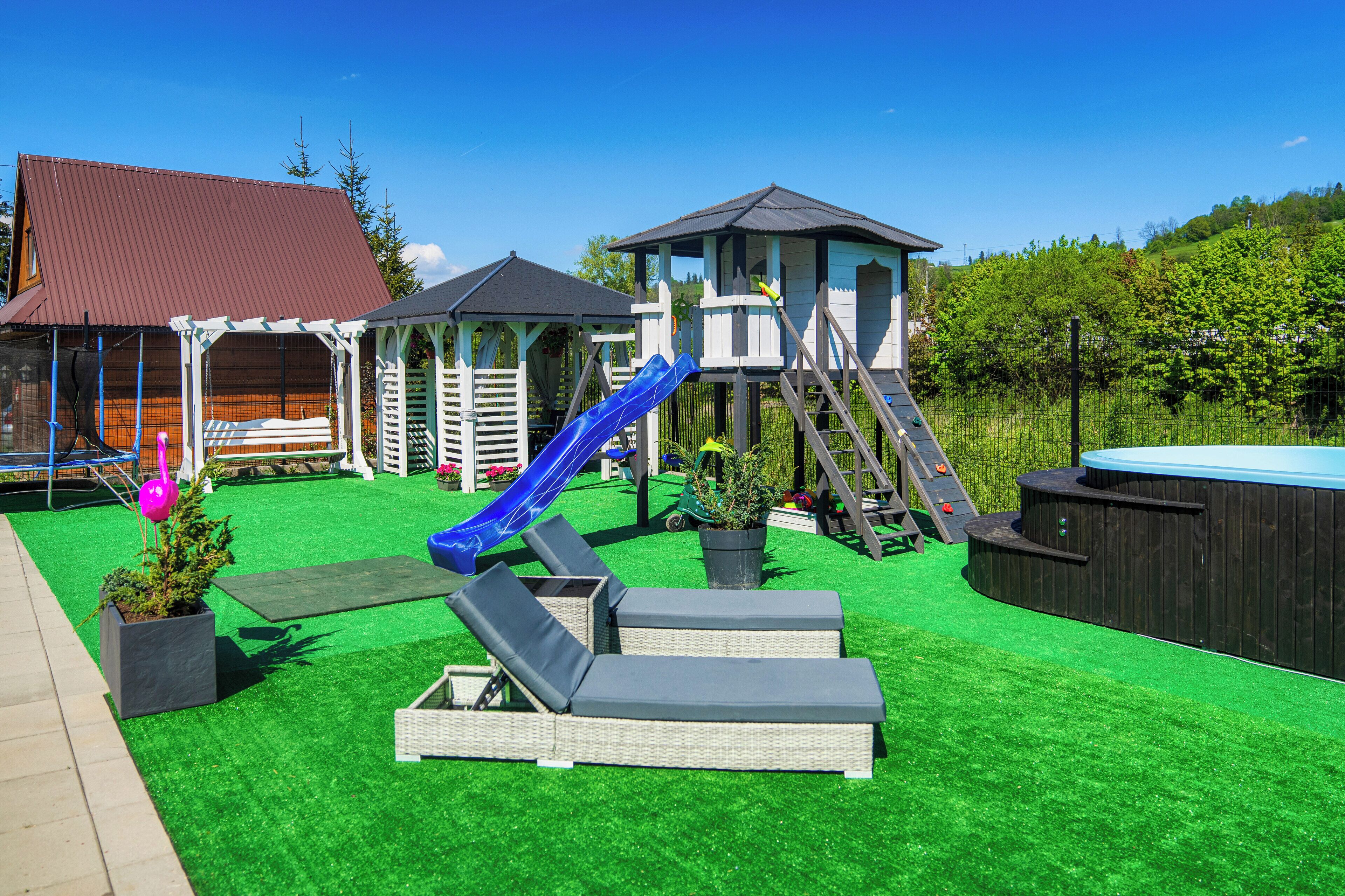 Un patio con césped artificial, un tobogán, sillas de descanso, un jacuzzi y una casita de juegos de madera.