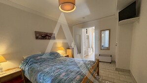 A bedroom with a bed, bedside table, lamp, and a small red chair. There is a door leading to another room and a mirror on the wall. - REZ DE CHAUSSEE, 1ERE LIGNE MER, ACCES DIRECT PLAGES (Sète)