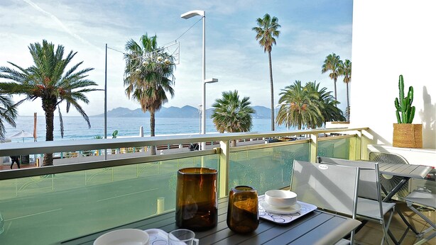 A balcony with a table set for dining, overlooking a coastal area with palm trees and mountains. - Apartment facing the beach (Cannes)