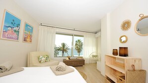 A hotel room with a bed, a wicker bench, a chair, a wooden shelf with decorative items, and a large window with curtains. - Apartment facing the beach (Cannes)