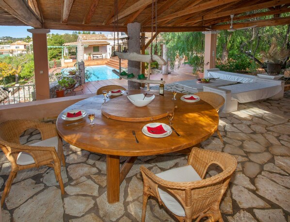 A wooden outdoor dining area with a stone floor, surrounded by wicker chairs and a white bench.