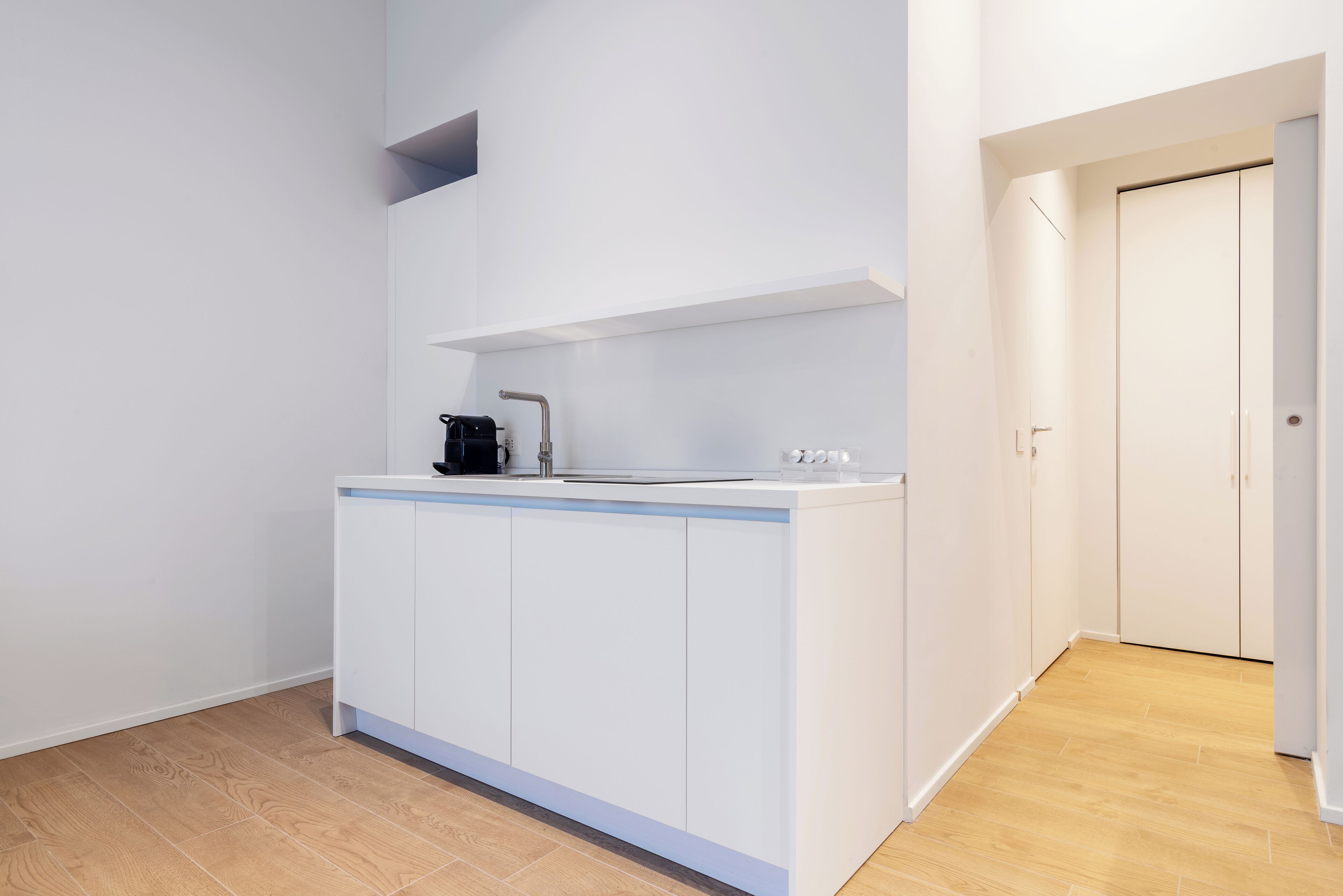 A modern kitchen with white cabinetry, a countertop, and a sink. There is a small shelf above the sink and a door to the right.