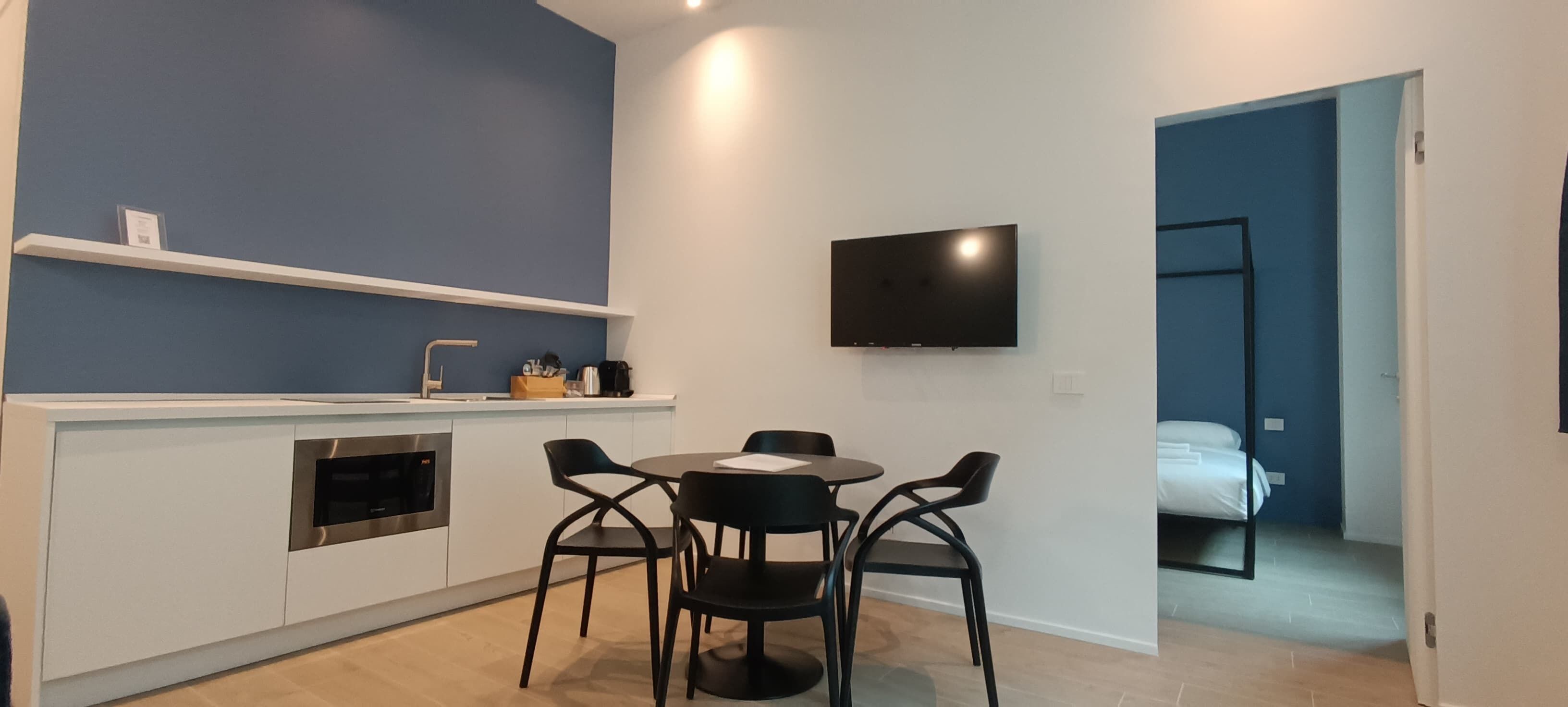 A modern kitchen with a built-in oven, a dining area with a round table and four chairs, a flat-screen TV mounted on the wall, and a doorway leading to a bedroom with a bed.