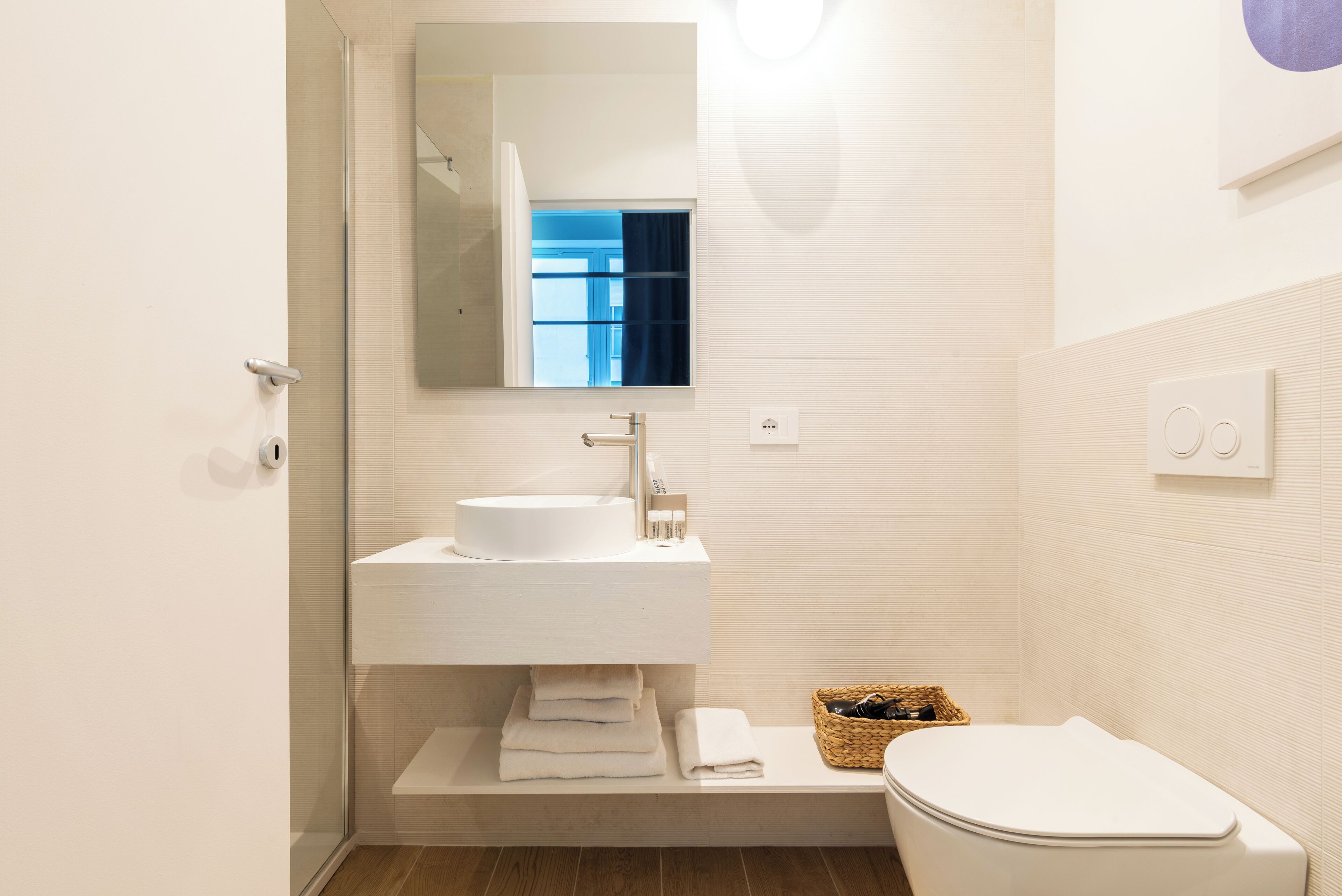 Un baño moderno con un lavamanos blanco, un espejo, un inodoro y una cesta de artículos de aseo.